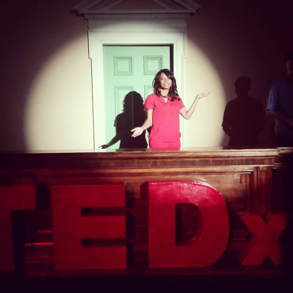 Antionette Kerr on stage under a spotlight, arms open toward the audience, with large red TEDx letters in front of the stage.